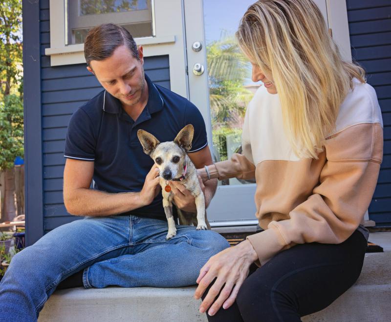 Two people sitting outside with a small dog on stairs leading into a hous